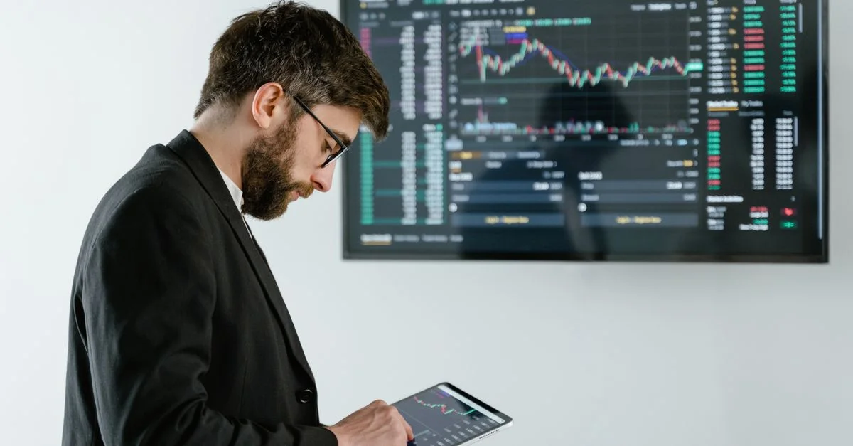 Homme professionnel utilisant une tablette devant un écran de trading de cryptomonnaie à l'intérieur.