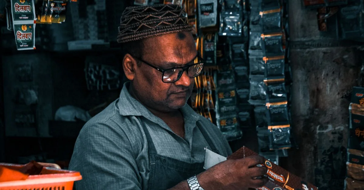 Un commerçant sud-asiatique mature organise des produits dans son magasin, portant des lunettes et un bonnet à motifs.