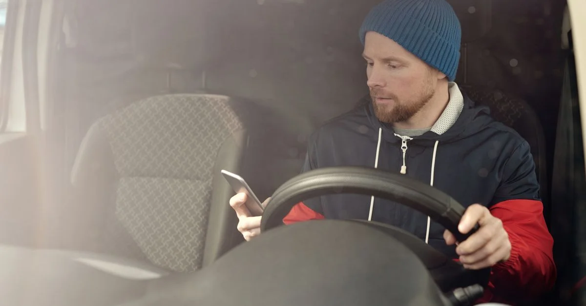 Un homme en bonnet utilise son smartphone tout en étant assis dans une voiture. En pleine lumière du jour.