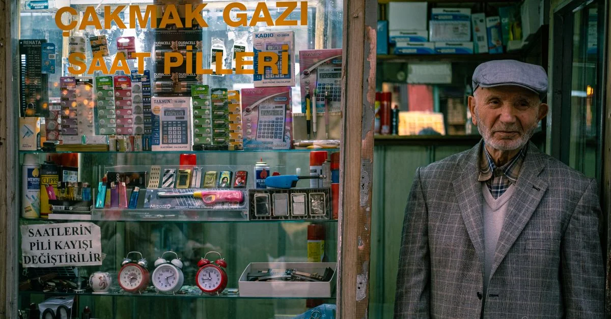 Un homme âgé se tient devant la vitrine d'un magasin d'électronique vintage exposant diverses piles et gadgets.