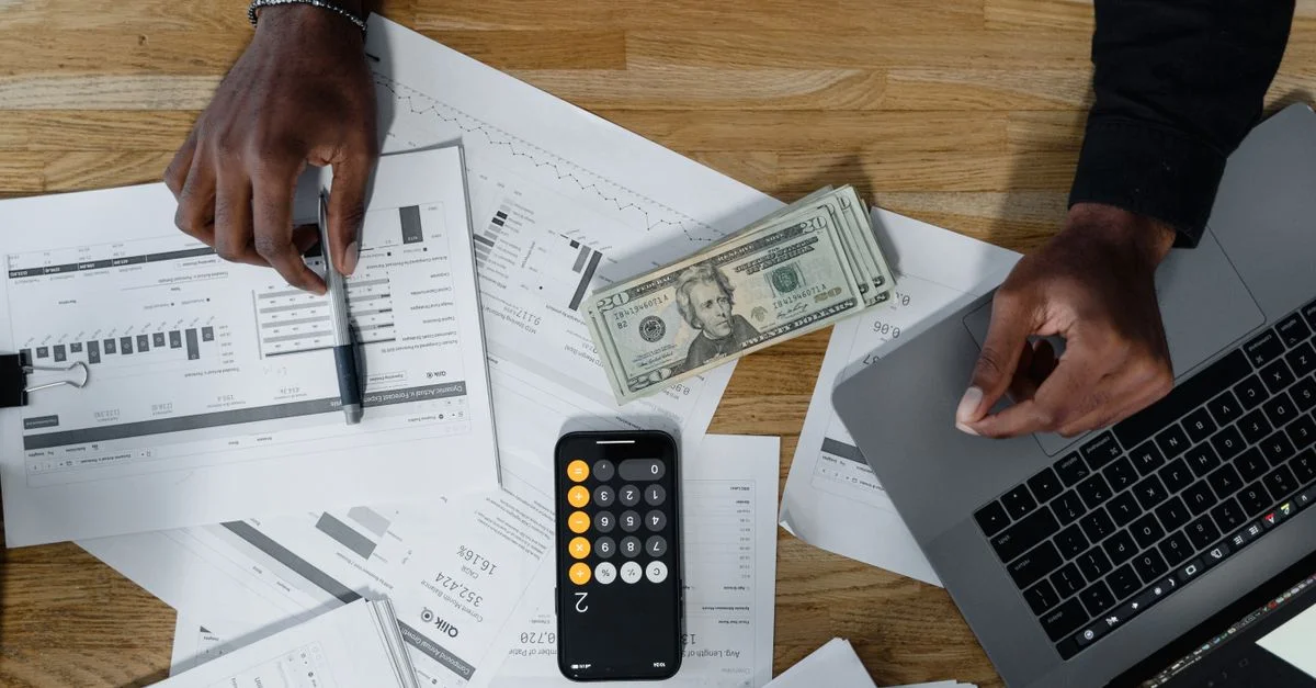 Des mains calculent des finances avec des papiers, des espèces et un ordinateur portable sur un bureau en bois.