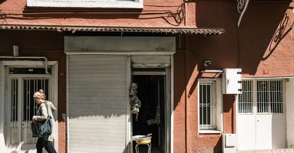 Une femme passe devant un magasin fermé par une journée ensoleillée, projetant des ombres sur le trottoir.
