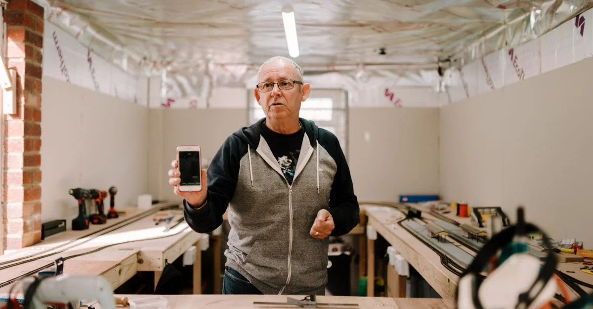 Un homme senior se tenant dans un atelier, montrant son smartphone avec une expression concentrée.